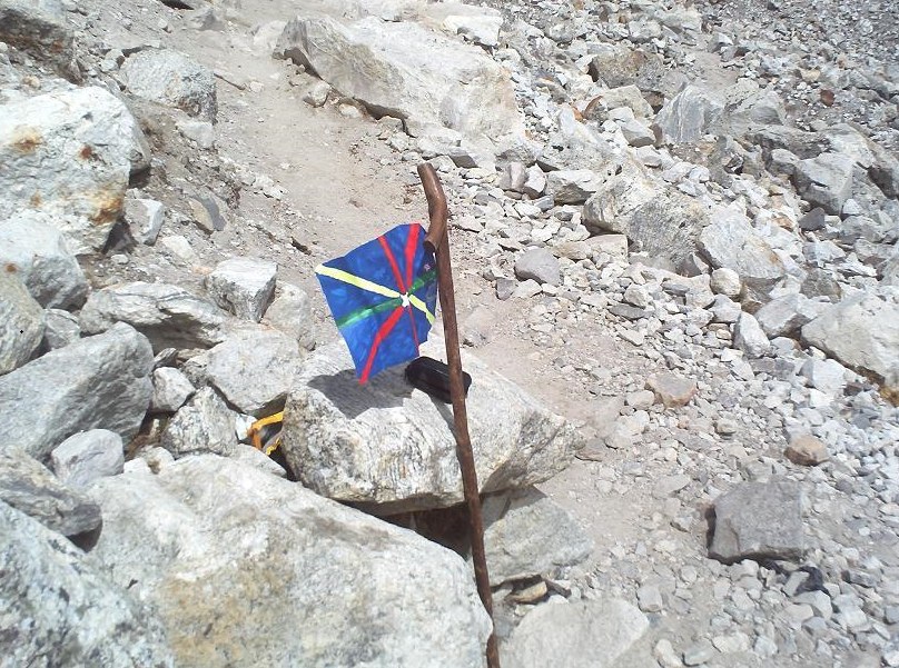 A rocky, boulder-strewn trail with a small blue flag with red and yellow stripes tied to a wooden walking stick